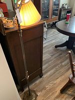 Full view of the vintage brass floor lamp standing next to a wooden cabinet and round dining table on a wood floor. The lamp is illuminated showing the cream-colored pleated fabric shade.