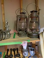 Two antique oil lanterns with rusted metal frames and glass chimneys, some accessories visible beside and below the shelf.