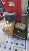Five vintage luggage pieces grouped on floor including a blue vanity case, two yellow suitcases, one beige and one dark brown suitcase.