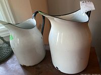 Two metal milk jugs on wooden surface, both white with black handles and trim. Visible signs of scratches and rust spots on both.