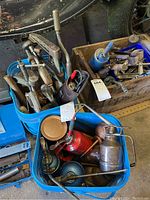 Overall view of two pails holding assorted metal oil cans and clamps with a wooden crate in background.