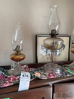 Two White Flame Light Company oil lamps on a floral runner, one with rounded amber font and clear base with handle, the other with amber font on tall pedestal base. Both have glass chimneys.