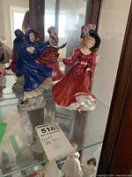 Two Royal Doulton figurines displayed in a glass cabinet: one in a red dress with black hat and the other in a blue dress with hood.