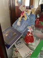 Two Royal Doulton porcelain figurines displayed in a glass cabinet alongside the Royal Doulton Collectors Record Book; one figurine in blue dress (8 inches), the other in red dress (4 inches).