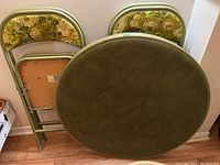 Full view of the folded round table showing the smooth olive green top and along with two folded chairs showing floral padded upholstery.
