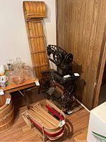 Photo showing the tall wooden toboggan leaning against wall next to a sewing machine and various glassware, with the small red sleigh on the floor in front.