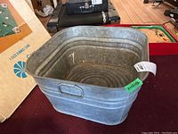 Photo showing side angle of weathered metal tub with handle, placed on carpeted floor with other items around.