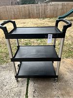 Full view of the three-tier black rolling utility cart outdoors on concrete with a wooden fence and playground slide in background.
