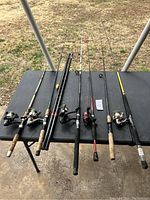 Six fishing poles with attached reels laid out on a black table, outdoors on grass background.