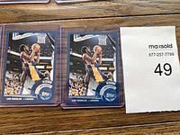 Two Kobe Bryant basketball cards in clear plastic sleeve on wooden surface with MaxSold bidding sheet