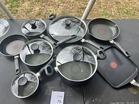 Overview photo showing various T-Fal cookware items including multiple frying pans, saucepans, sauté pan, grill pan and lids arranged on a black surface outdoors.