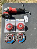 Milwaukee angle grinder with two Norton nylon disc brush attachments still in packaging and two CGW abrasive flap discs laid out on table.
