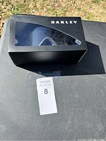Image of unopened black Oakley box with clear window showing black snow goggles inside, placed on outdoor surface with auction lot tag visible.
