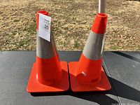 Two orange traffic cones side by side on a black table outdoors, showing their full height and square bases.