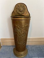 Frontal image of antique brass umbrella stand showing detailed embossed tavern scene with seated figure and dog drinking from bowl at bottom.