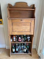 Front view of antique secretary desk with drop front writing surface and shelves below holding liquor bottles, showing wood grain and drop front secured by lock.