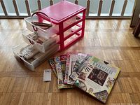 Pink plastic 4-drawer storage unit with clear drawers, containing embroidery threads and scissors, with an embroidery hoop resting on top, sitting on wooden floor.