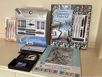 Photo showing various calligraphy kits and stationery including Sheaffer calligraphy set, The Lettering Calligraphy Project Kit, pastel cardstock pack, ink bottles, and pens.