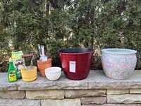 Wide view of all items arranged on stone wall showing two large planters on right, three smaller pots and tools on left, and Miracle-Gro plant food bottle on far left.