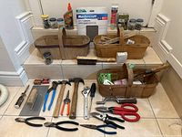Photo showing two woven baskets, bottles of glue, hardware boxes, wire brush, multiple tool types including hammers, pliers, and screwdrivers arranged on a tiled floor with tiled steps behind.