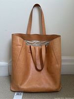 Front view of tan leather tote bag with two long shoulder straps, standing upright showing natural creasing and wear.