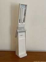 White OttoLite desk lamp angled showing compact fluorescent bulb, adjustable hinge, on wooden surface against white wall.