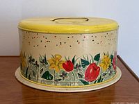 Photo of the vintage floral cake tin showing yellow lid with metal handle and floral graphics with paint wear and spots.