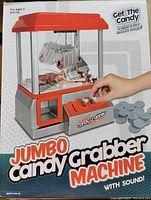 Front view of the Jumbo Candy Grabber Machine showing the red top, clear enclosure, claw mechanism, coin slot and container, and included plastic coins.