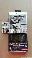 Photo showing three boxed items in lot: mirror mount phone holder, night driving glasses, silicone pop stand.