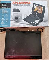Sylvania 7-inch Portable DVD Player box and player showing the device closed with power cord visible.