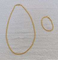 Full view of gold tone rope link chain necklace and matching bracelet laid on a light textured surface.