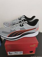 Side angle view of gray Puma running shoes on top of red Puma shoe box, showing black and orange logo detail.