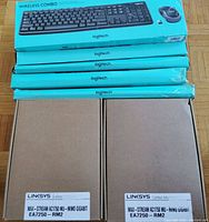 Five Logitech wireless keyboard and mouse combos in original teal Logitech boxes and four brown Linksys boxes labeled Max-Stream AC1750 MU-MIMO Gigabit EA7250-RM2.