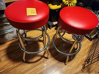 Two chrome bar stools with round red upholstered seats on wooden floor, seller tag on top