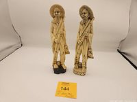 Two vintage Japanese resin figures of men with sticks and ducks on their backs, side by side showing front view