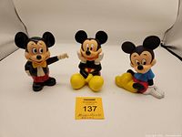 Front view of three Mickey Mouse coin banks showing different poses and colorful outfits.