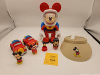 Shows all Mickey Mouse themed items including two Fisher-Price Little People cars with Mickey and Minnie figures, large Mighty Bean astronaut figure, small figures, and the beige visor hat with Mickey embroidery.