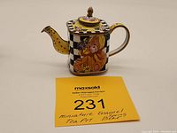Front and side view of a miniature enamel teapot with a teddy bear wearing a bonnet and a checkerboard design