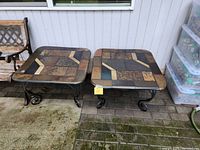 Photo displaying two square slate tile patio tables with metal scroll legs positioned against a house wall and patio bricks beneath. Tables appear wet with weather wear.