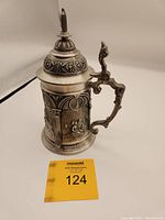 Front and side view of vintage pewter German beer stein showing detailed embossed relief and ornate handle and lid.