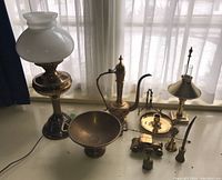 Wide view of lot on white surface showing brass lamp with milk glass shade, etched brass tea pot, pedestal bowl, brass car figurine, candlestick and hand bells
