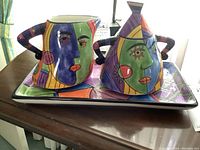 Front angle photo showing the ceramic cream pitcher and covered sugar bowl on the matching rectangular tray with colorful abstract faces.