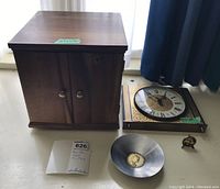 Photo showing closed wooden Olympic coin box, wall clock, Italian stainless steel coin dish, and miniature plate with farming scene arranged on a surface.