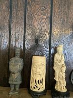 Three Asian-style figures including a resin Terracotta Army soldier and two cream-colored resin figures against wooden background.