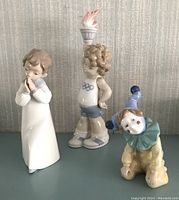 Three Lladro figurines displayed together: praying boy, Olympic torch bearer, and jester clown.