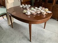 Full view of oval wooden dining table with warm medium brown finish. The table has tapered legs and is shown with a polished surface. Several small teacups and saucers are placed on the table for scale but not included.