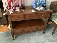 Full front view of the McLagan wood server with two drop leaves down, showing three small drawers and open shelf below with lower drawer, all resting on brass caster wheels.