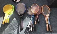 Lot showing yellow plastic badminton racquets, two black Cooper tennis racquets with metal frames, orange badminton racquets, and wooden Wightman tennis racquets on carpet.