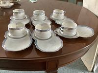 Six porcelain tea cups paired with matching snack plates, all white with silver trim, arranged on wooden table.