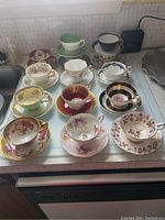 Photo showing 11 assorted teacups and saucers with various floral patterns and colors including green, pink, black, maroon, blue bases with gold trim and decoration.